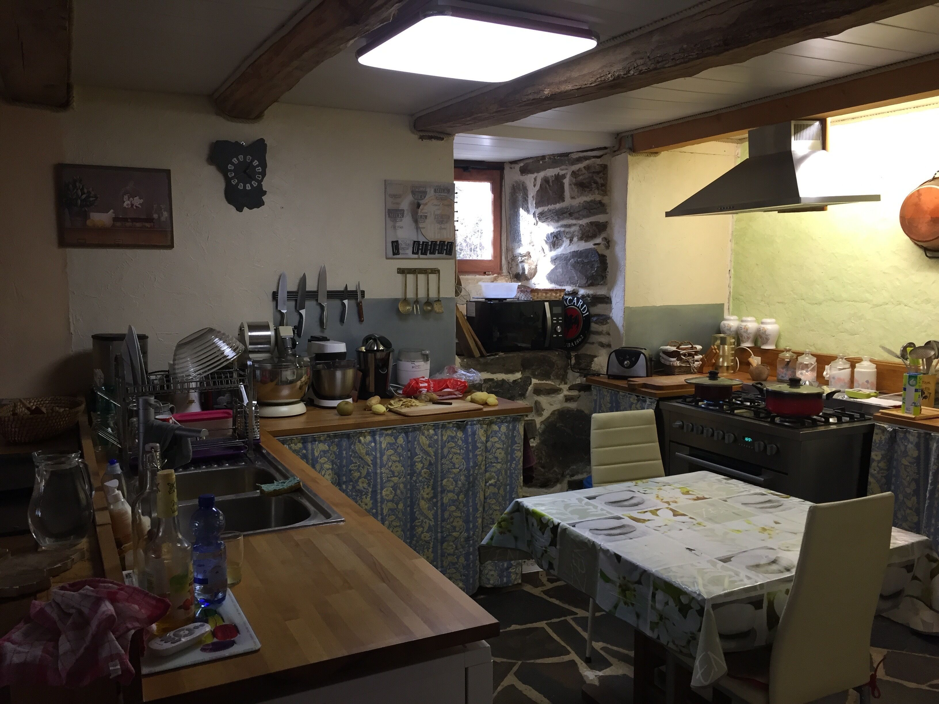 A kitchen with a stone wall, wooden beams, a gas stove, a sink, and a dining table with chairs.