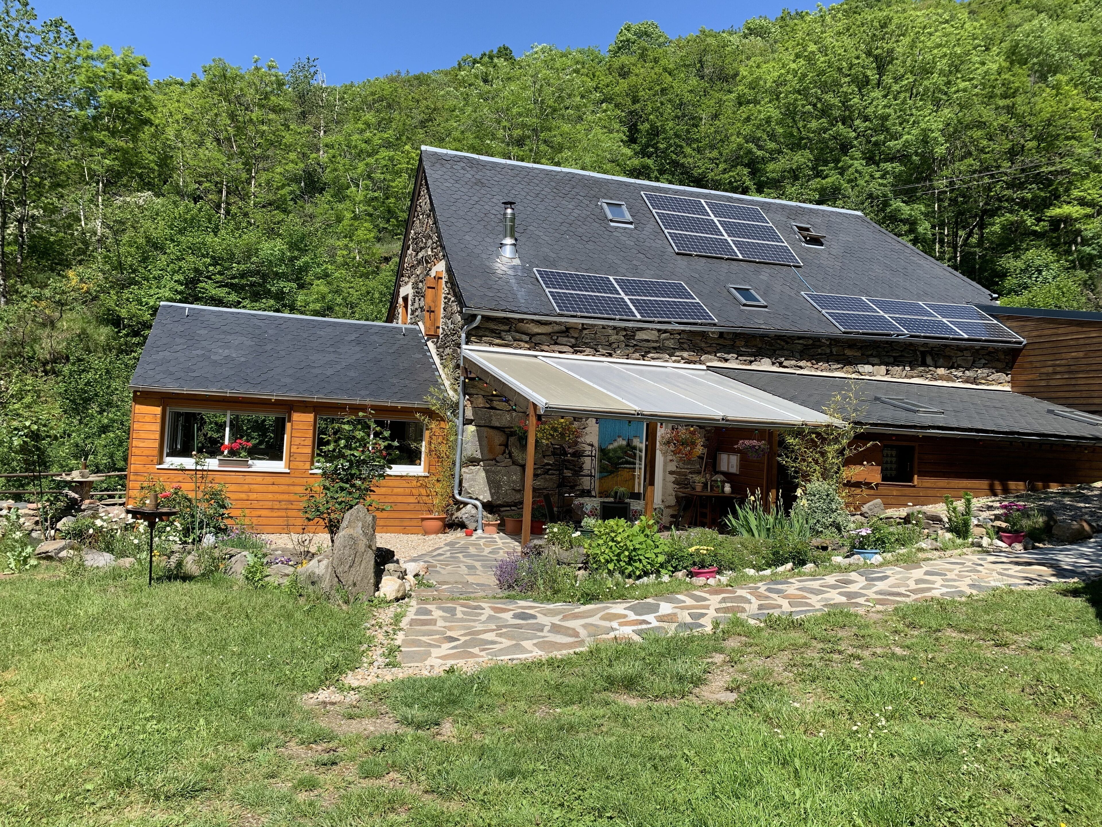 Une maison en bois à deux étages, avec des fondations en pierre, des panneaux solaires sur le toit et un chemin de pierre menant à l’entrée.