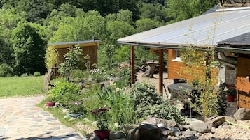 Een rustieke hut met een pad van stenen naar een houten terras, omgeven door weelderige begroeiing en diverse potplanten.