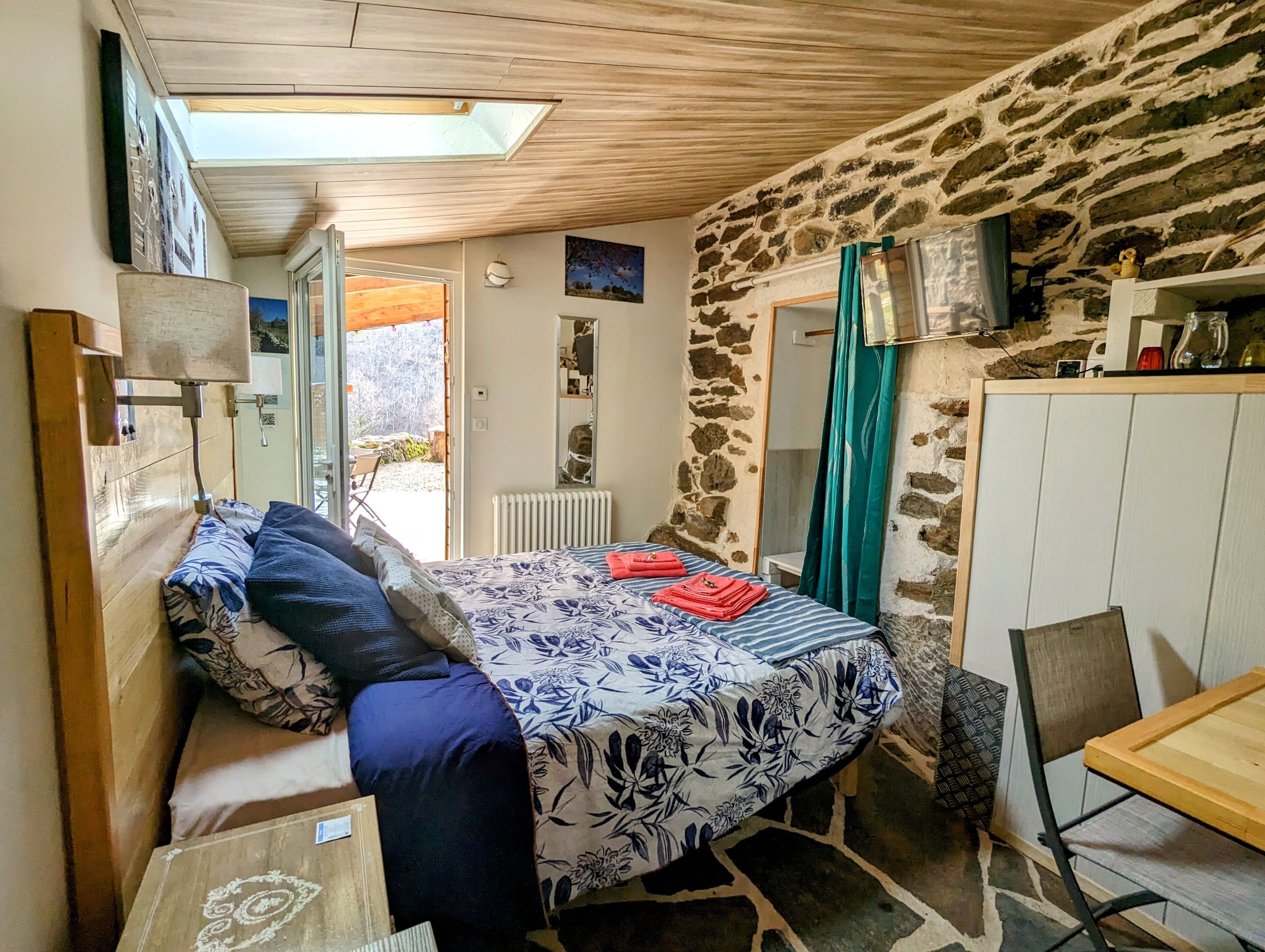 A cozy bedroom with a bed, a small table, a chair, a radiator, and a stone wall.