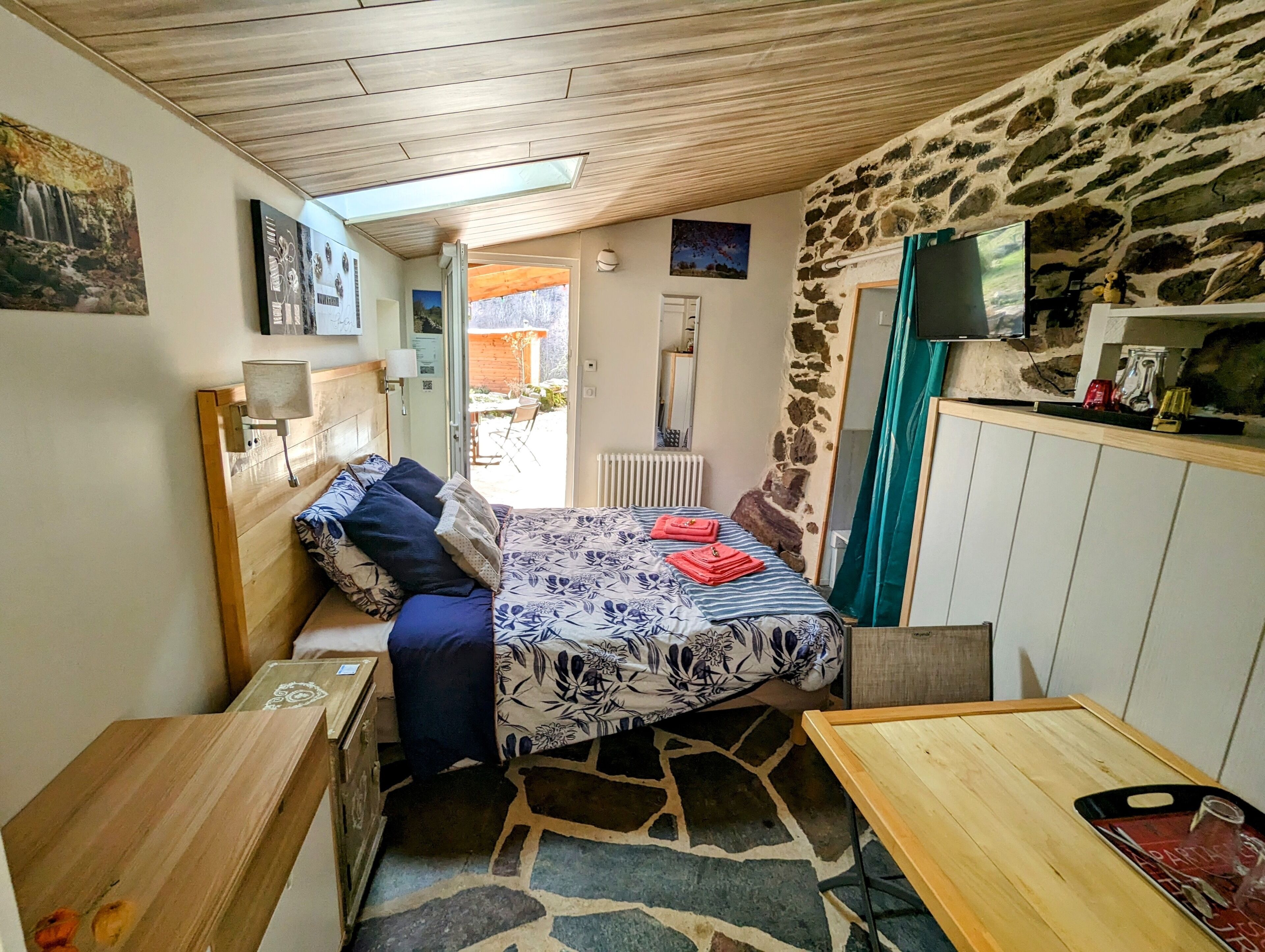 A compact room with a bed, a small table, a TV, and a stone wall.