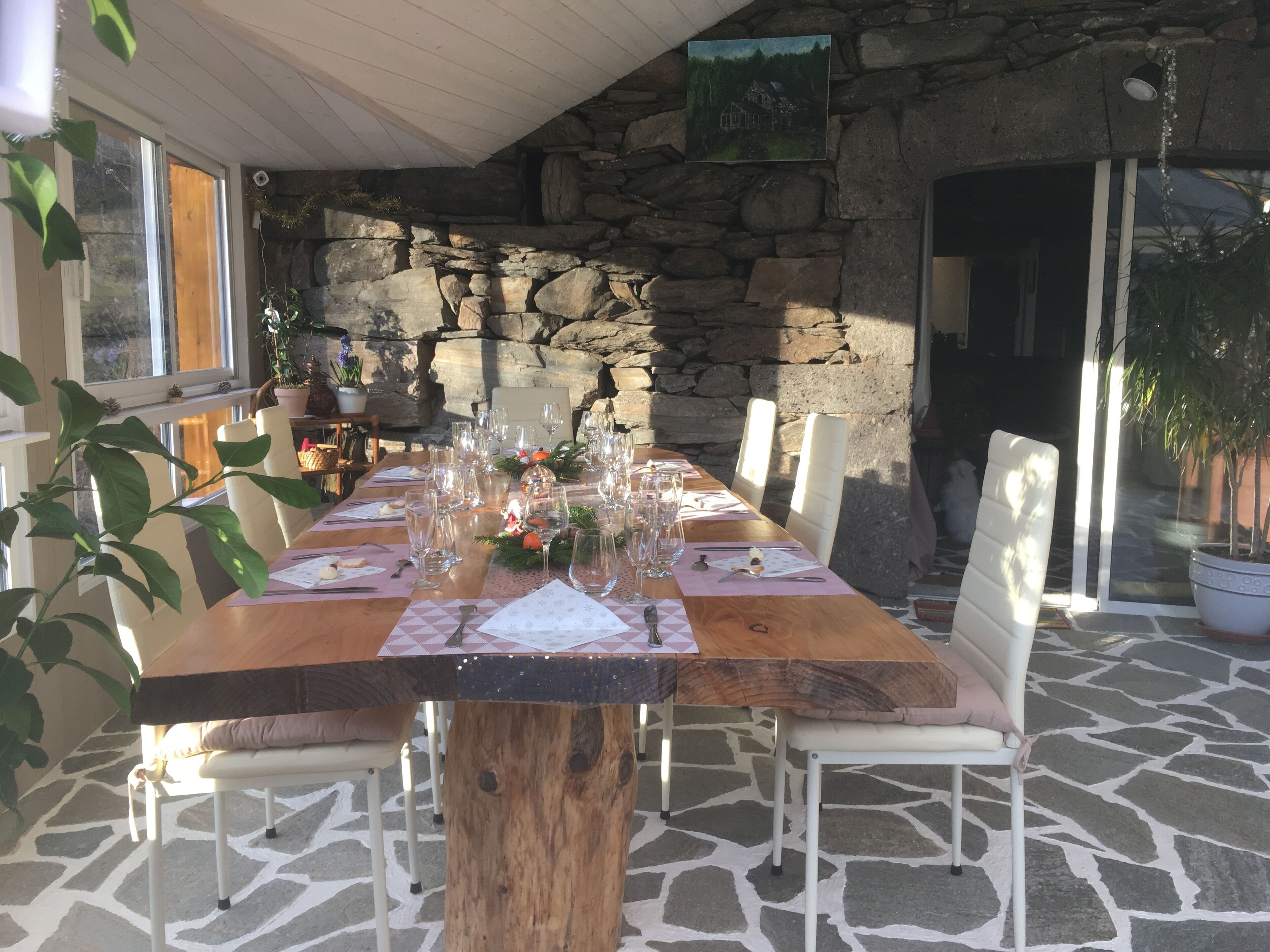 Un tavolo da pranzo in legno apparecchiato per sei persone, con sedie bianche, circondato da pareti in pietra e piante in vaso.