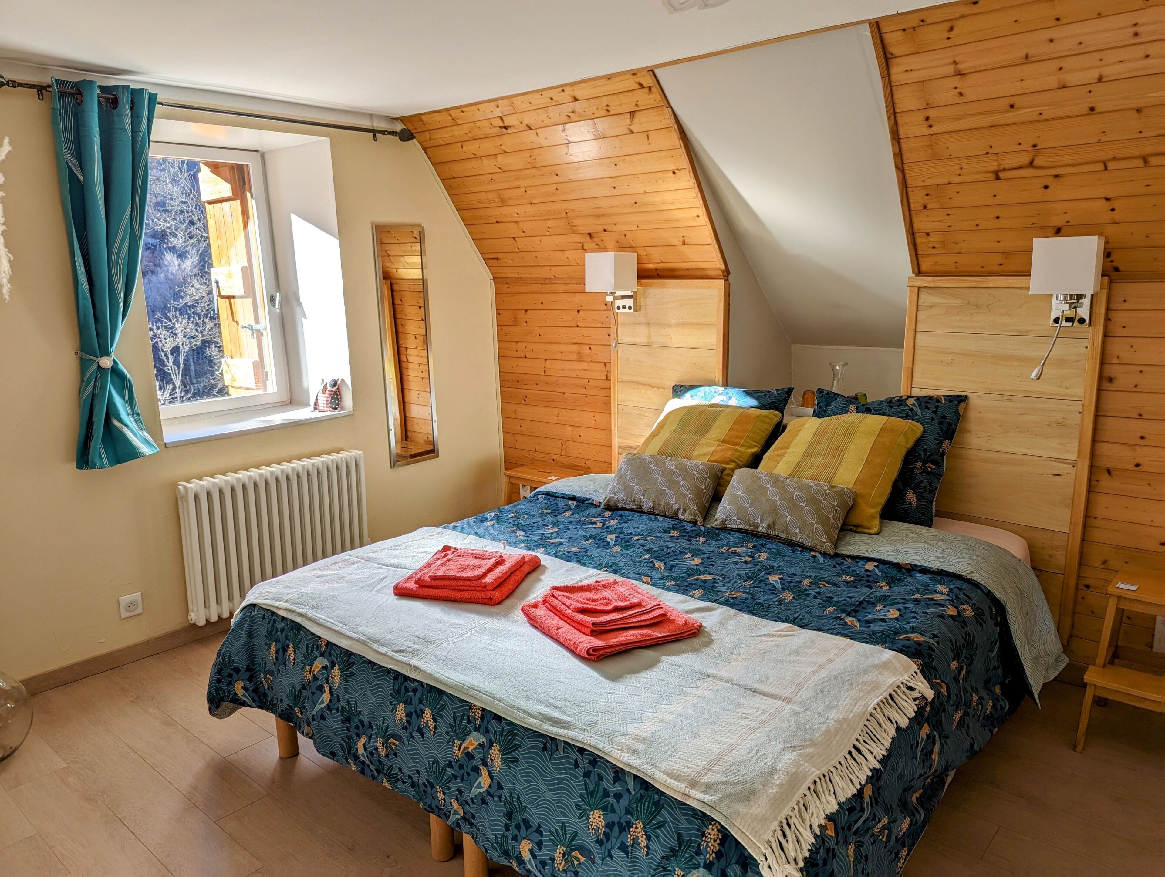 A bedroom with a sloped ceiling, wooden walls, a bed with multiple pillows, a window with curtains, and a radiator.