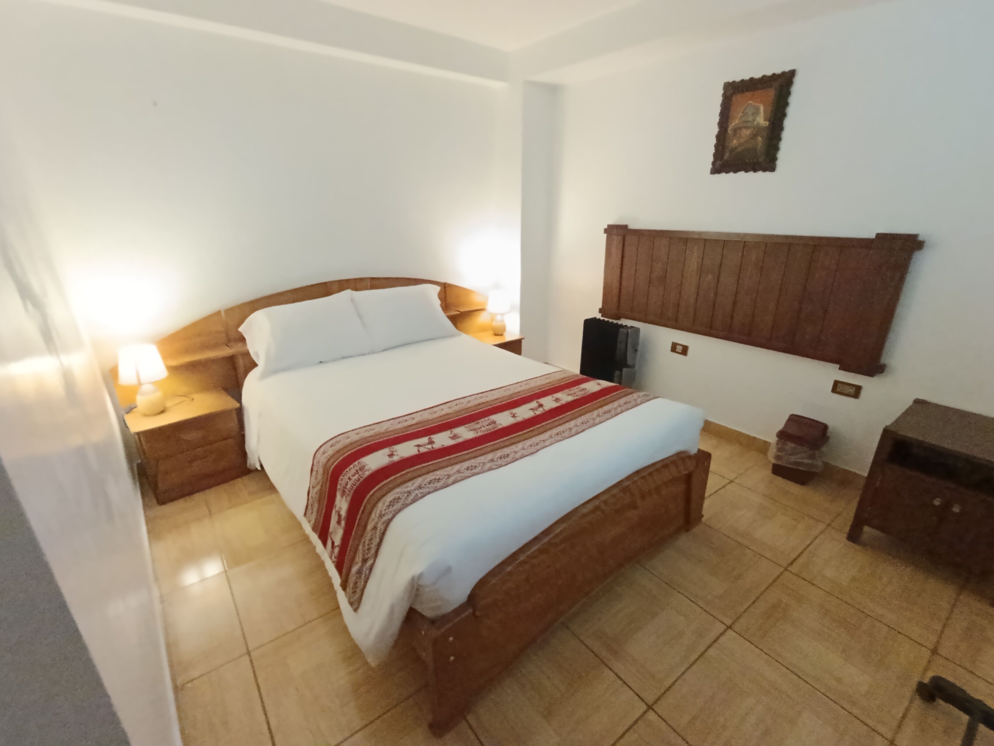 Een slaapkamer met een houten bed, wit beddengoed, een nachtkastje met lamp, een houten hoofdbord, een klein houten kastje en een wandplank.