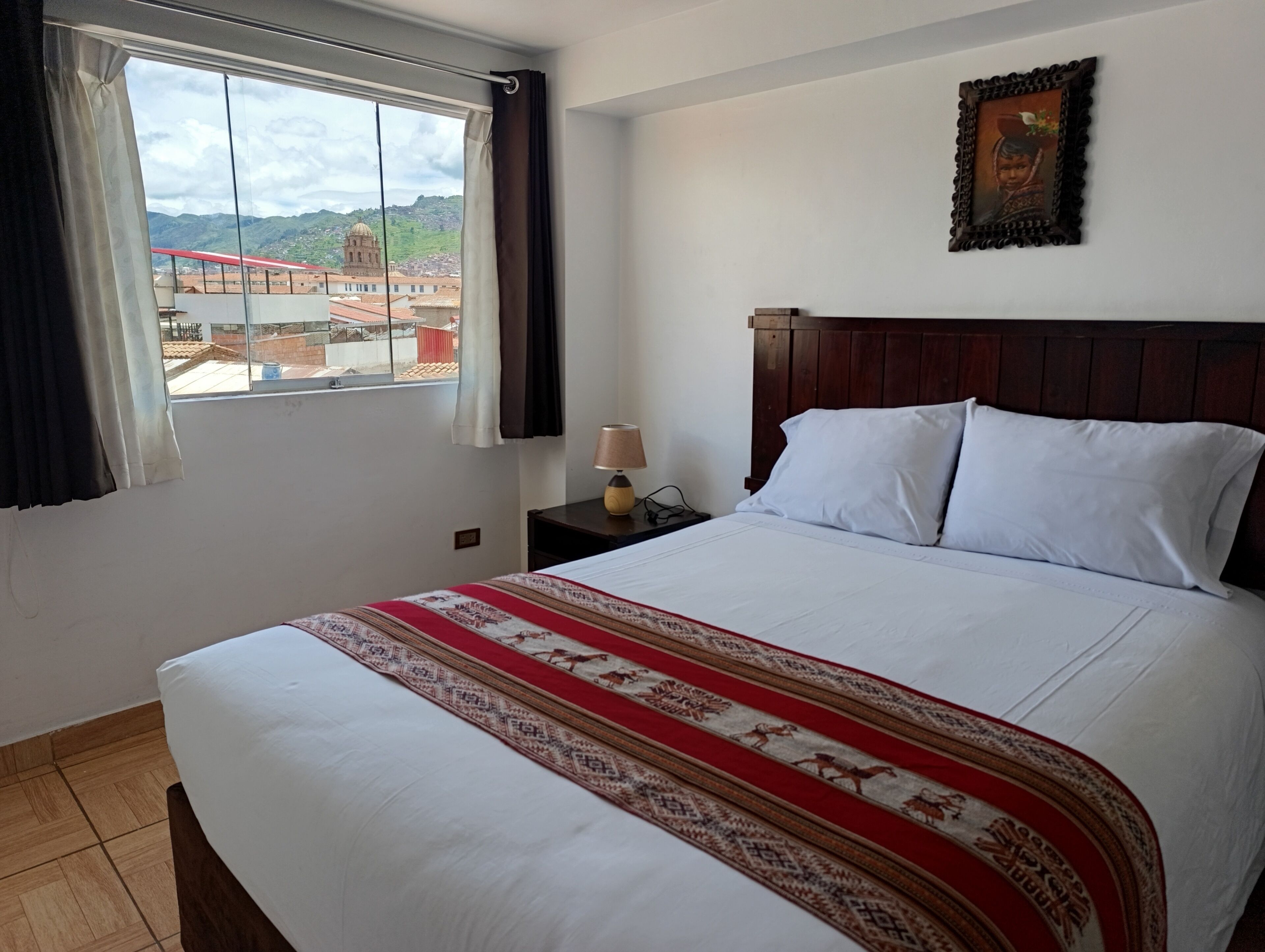 A hotel room with a large bed, a wooden headboard, a nightstand with a lamp, a window with curtains, and a framed picture on the wall.