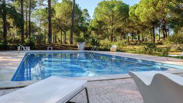 A rectangular swimming pool with a tiled edge, surrounded by a concrete deck. There are two white lounge chairs and a white outdoor chair near the pool.