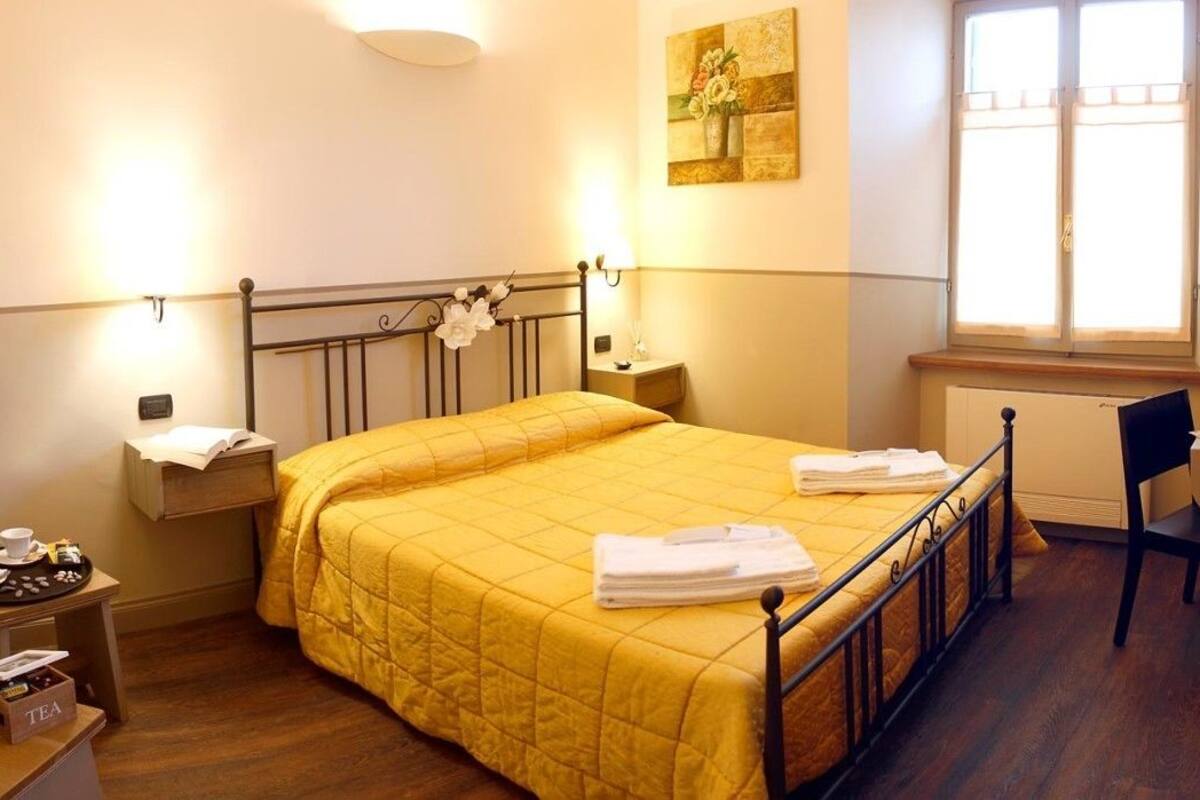 A hotel room with a bed, bedside tables, a chair, a window, and a painting on the wall.