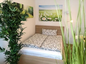 Un dormitorio con una cama, una planta verde grande y dos cuadros en la pared.