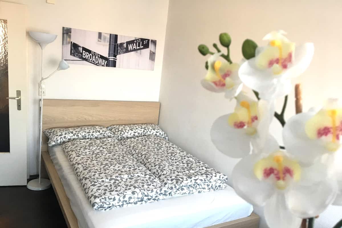 Un lit bien fait, avec une literie à motifs, une lampe de chevet, un panneau de rue encadré et une plante d’orchidée blanche.