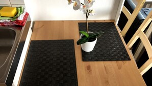 A wooden table with two black woven placemats, a white pot with a green plant, and a vase with white flowers. - Downtown (Regensburg)