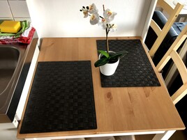 Una mesa de madera con dos individuales negros tejidos, una maceta blanca con una planta verde y un florero con flores blancas.