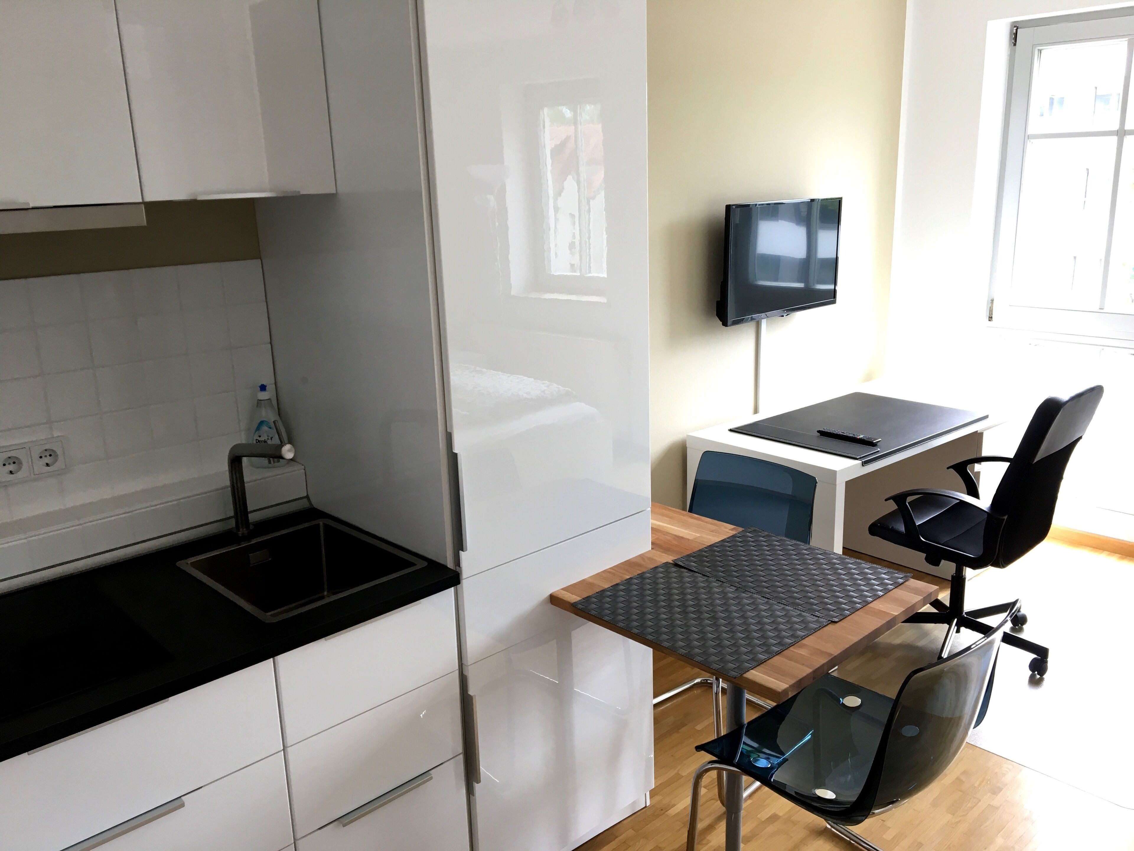 Une cuisine moderne avec des meubles blancs, un plan de travail noir et un lave-vaisselle encastré. On y trouve une petite table avec une chaise noire et un téléviseur fixé au mur.