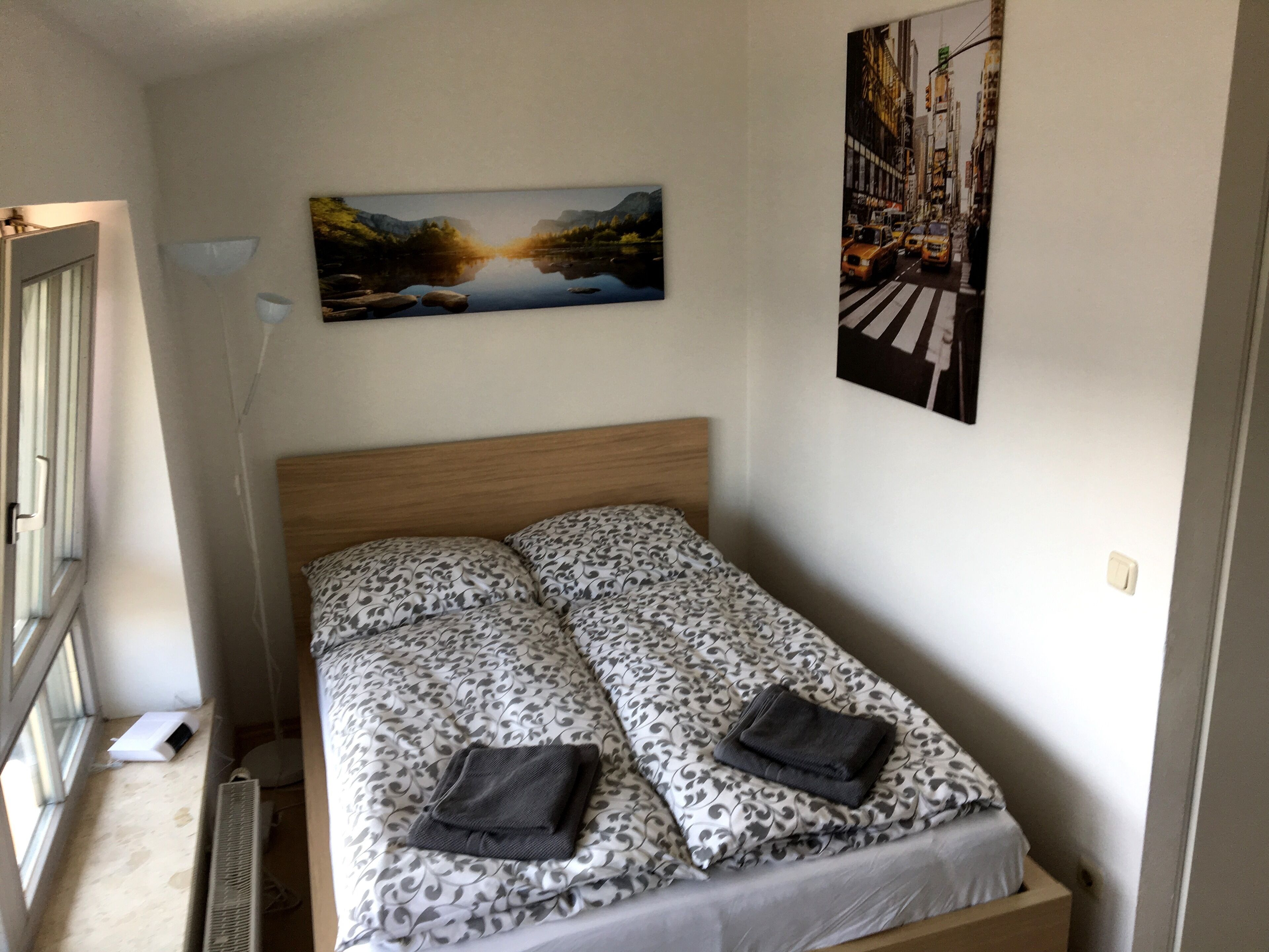Ein Einzelbett mit gemustertem Überwurf, einem Kopfteil und zwei Kissen. Links befindet sich ein Fenster und ein Bild hängt über dem Bett.
