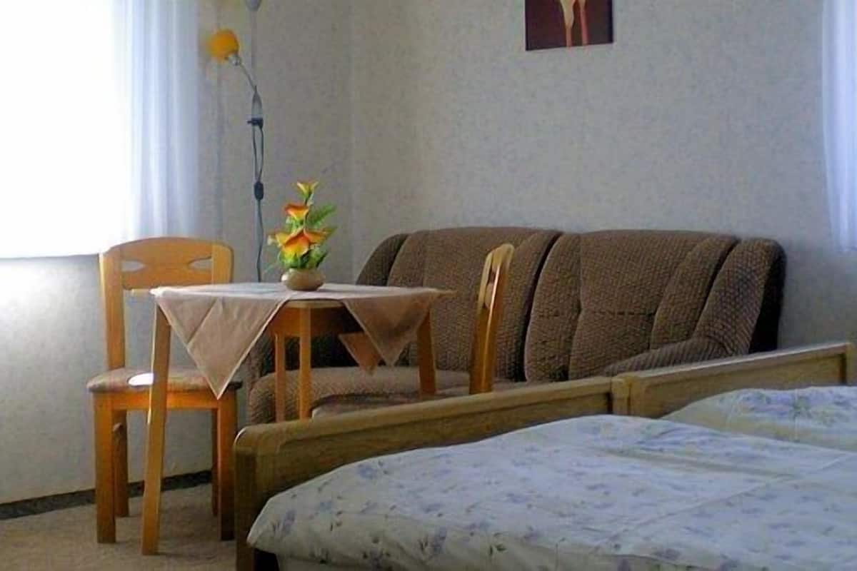 A bedroom with a bed, a chair, a small table with a vase of flowers, and a window with curtains.
