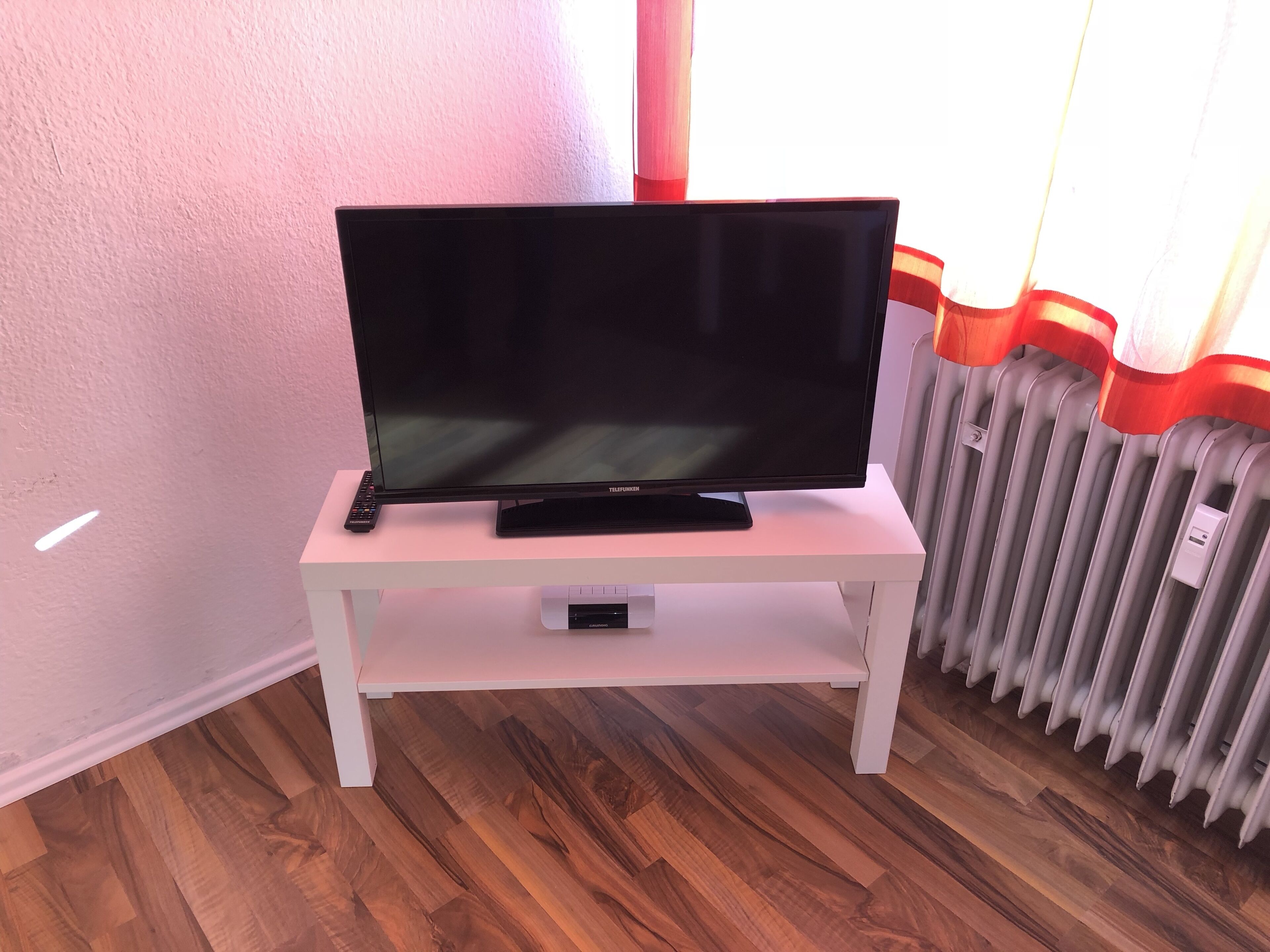 Ein Flachbildfernseher auf einem weißen TV-Schrank in einem Zimmer mit Holzböden und einer Heizung.
