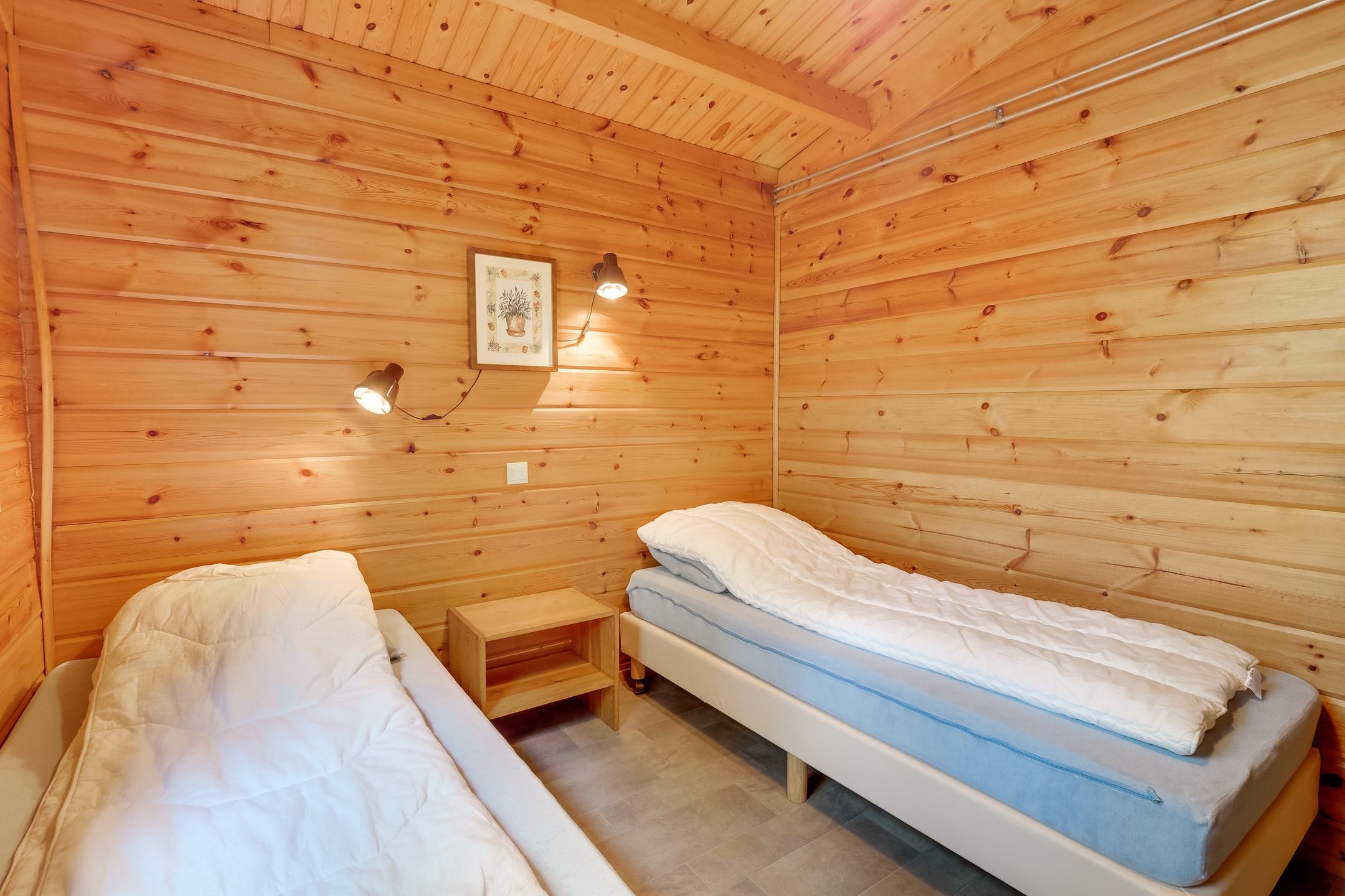 A wooden cabin room with two single beds, a small wooden nightstand, and a wall-mounted light fixture.
