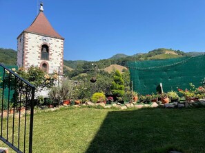 A tower with a clock, surrounded by a well-maintained garden with various plants and a clear view of the hills in the background.