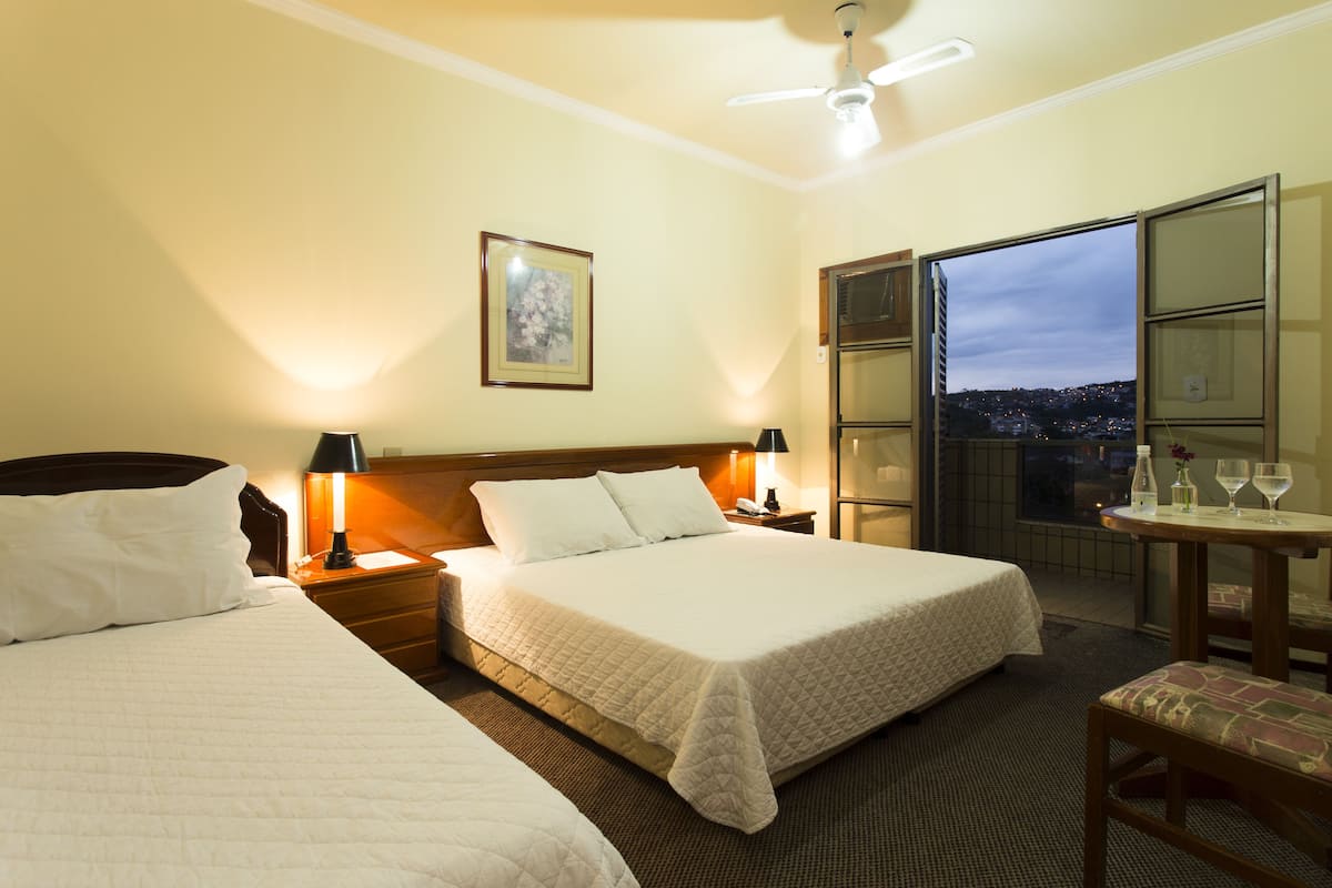 Ein Hotelzimmer mit zwei Betten, einem Balkon mit Blick auf die Stadt, einem Tisch mit Flasche und Gläsern sowie einem Gemälde an der Wand.