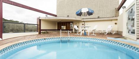 Une piscine avec une terrasse carrelée, un espace de détente couvert avec des chaises, et un bâtiment en arrière-plan.