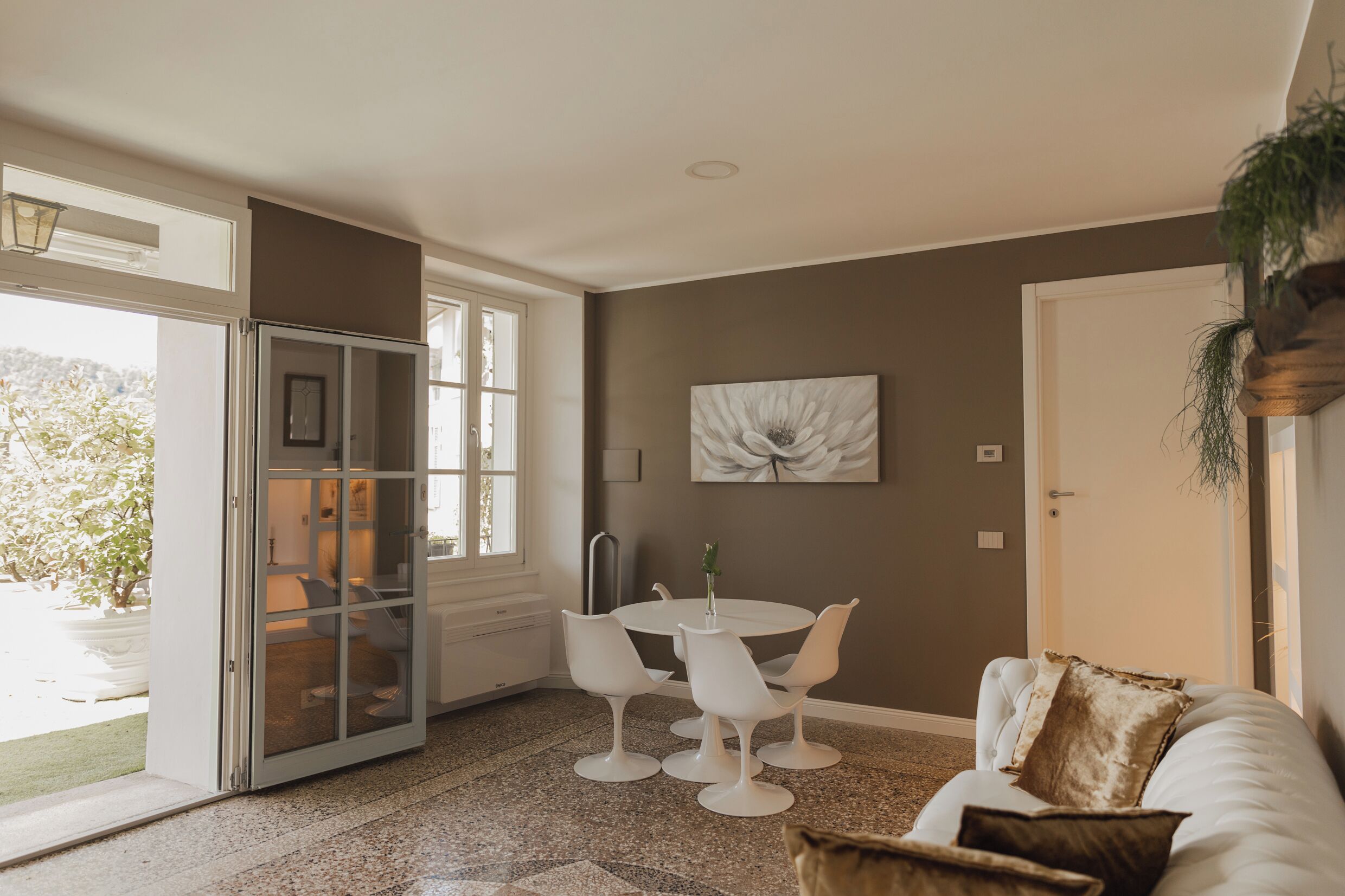 A room with a sliding glass door leading to a balcony, a round dining table with white chairs, a white sofa with a brown cushion, and a large abstract painting on the wall.