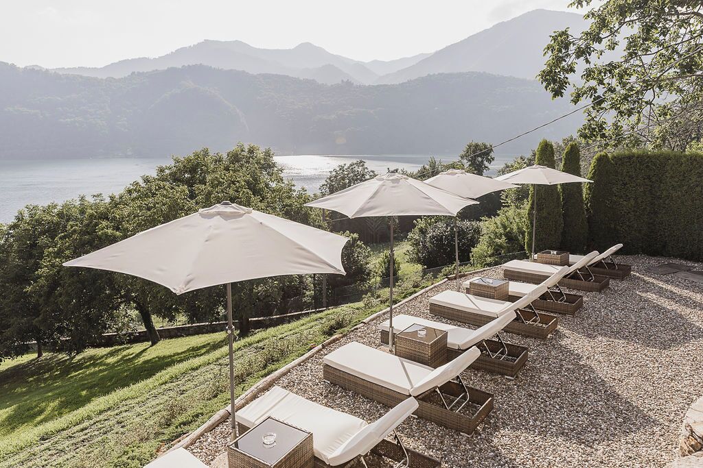 A row of sun loungers with umbrellas, a gravel path, and a view of a lake with mountains in the background.