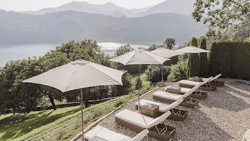 A row of sun loungers with umbrellas, a gravel path, and a view of a lake with mountains in the background.