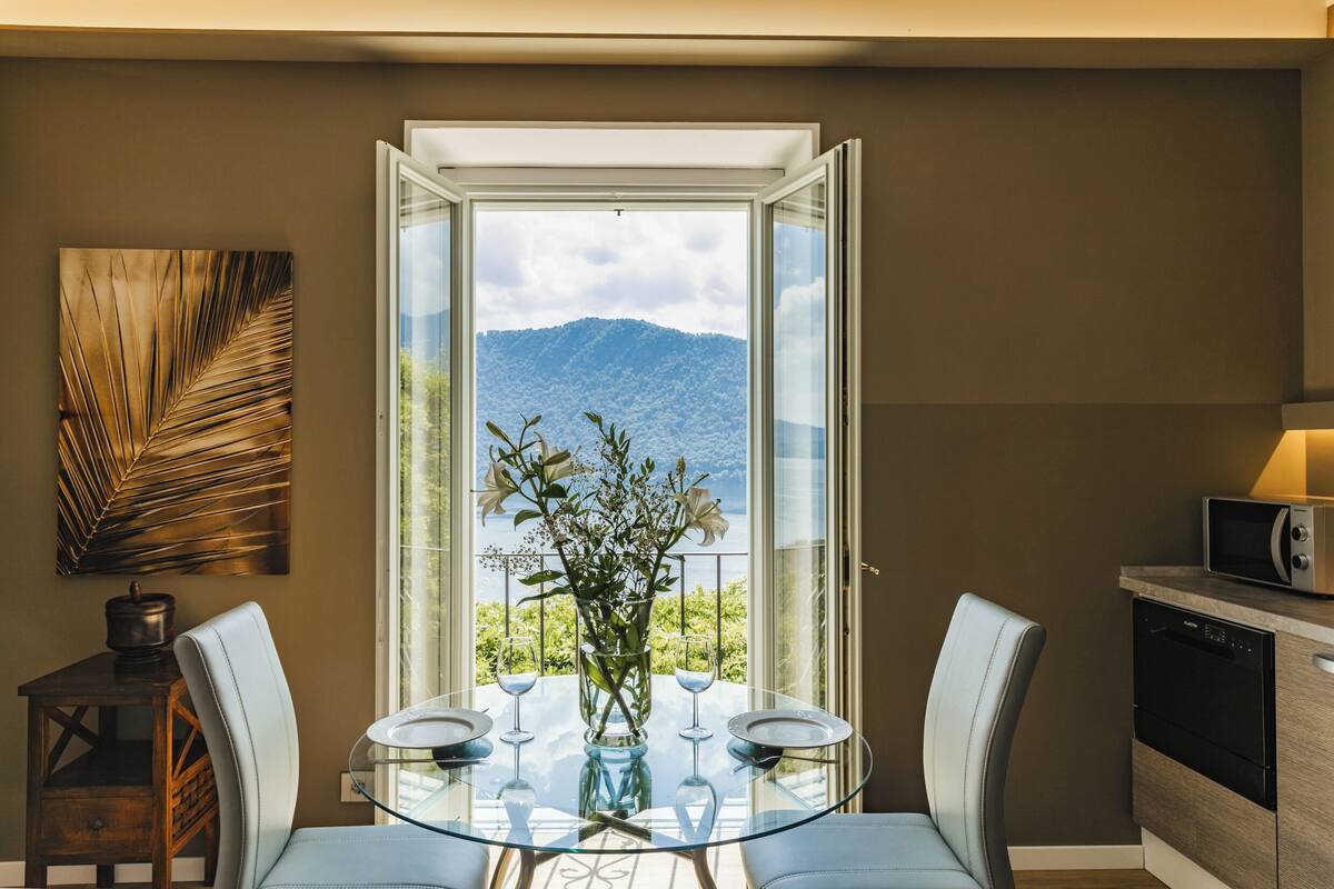 A dining area with a glass table, white chairs, a vase with flowers, and a view of mountains through a large window.