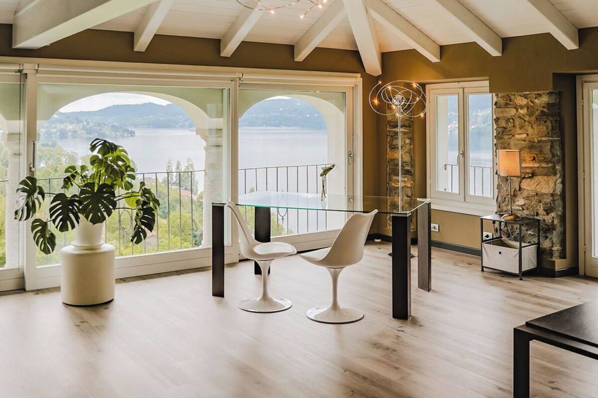 A spacious room with a high ceiling, large windows offering a view of a lake, a glass table with modern chairs, and a stone fireplace.