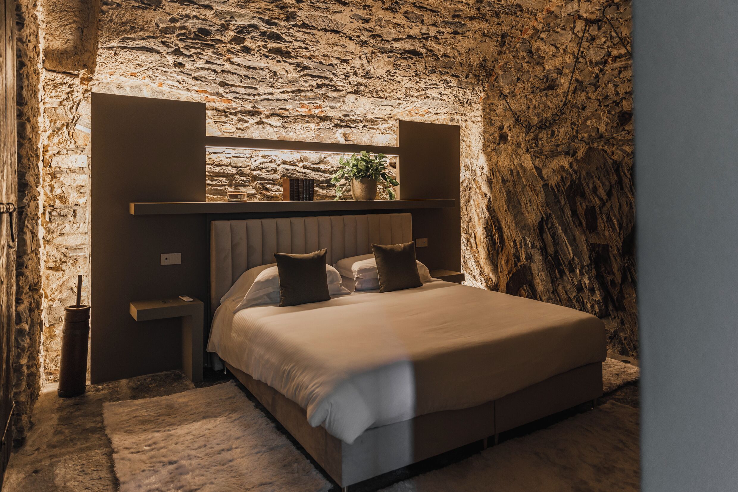 Un dormitorio con pared de piedra, una cama con cabecera, mesitas de noche y una planta en maceta.