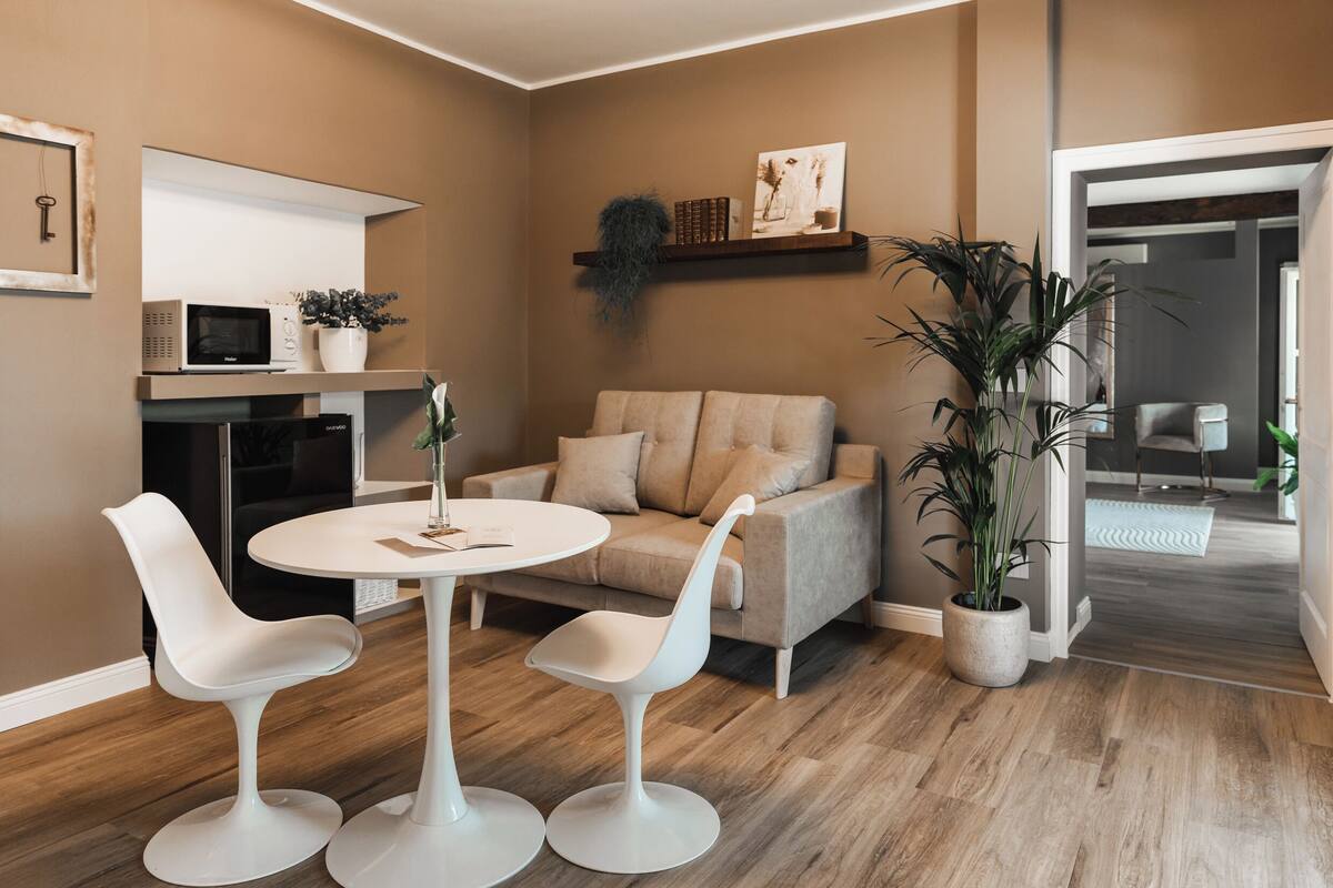 A modern living room with a beige sofa, white chairs, a round table, and a kitchen area with a microwave and potted plants.