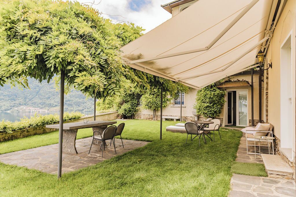 A patio with a stone table and chairs, a pergola with climbing plants, and a view of a lake.