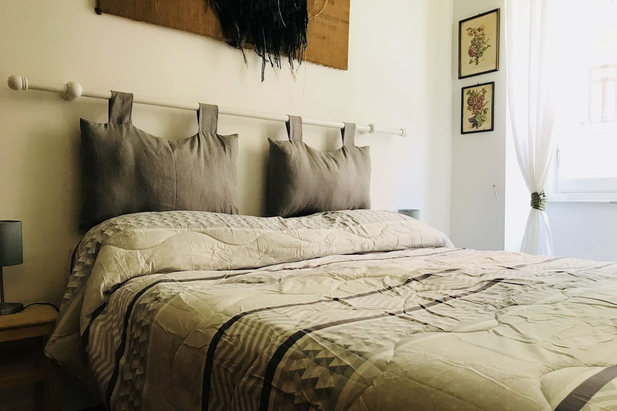 A neatly made bed with a patterned bedspread and pillows, a bedside table with a lamp, and a wall with a tapestry and framed pictures.