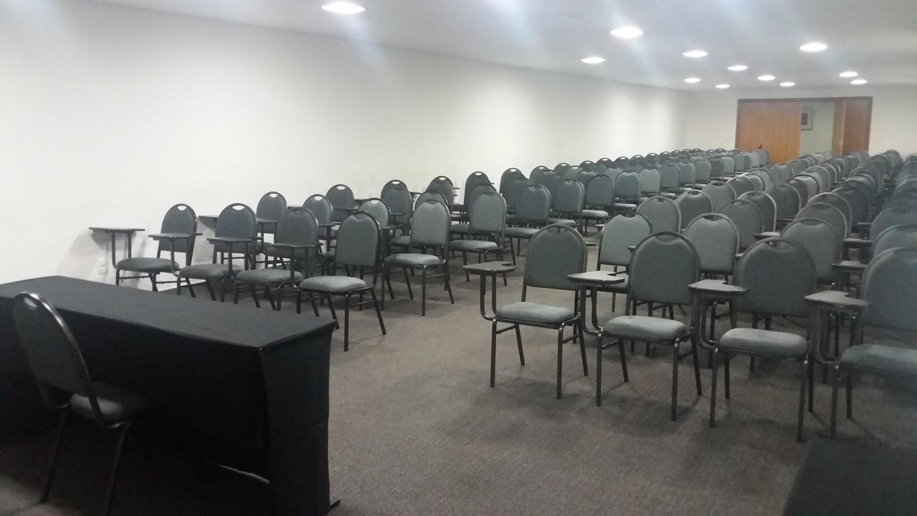 Un salón de conferencias con filas de sillas y mesas, una pantalla de proyector y una puerta al fondo.