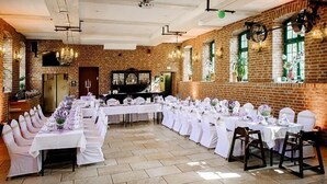 Un salón de banquetes con largas mesas dispuestas para un evento formal, paredes de ladrillo y ventanas amplias.