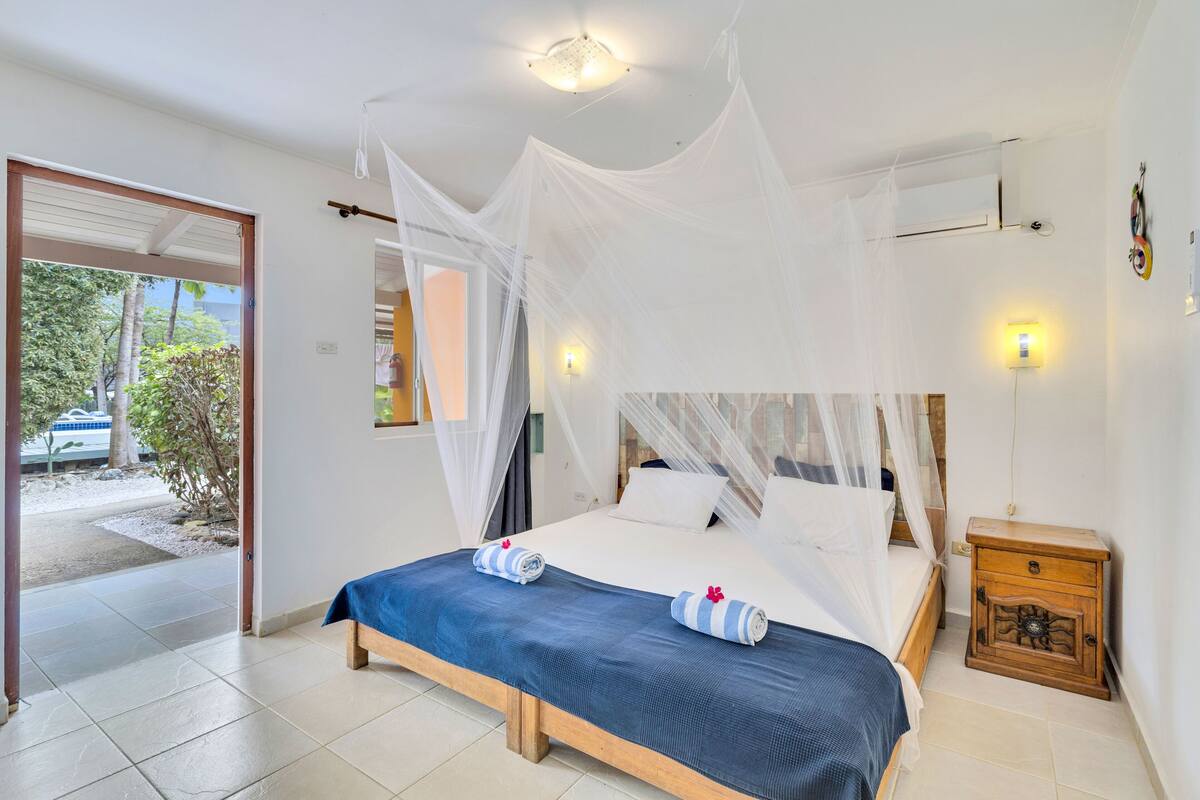 A bedroom with a bed, a nightstand, a window with curtains, and a door leading outside.