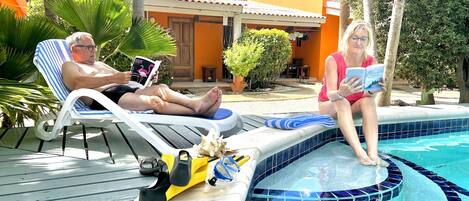 A poolside area with lounge chairs, a pool with blue mosaic tiles, and a building with orange walls and a tiled roof.