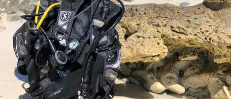 A black scuba diving backpack with various attachments on a sandy beach with rocks.