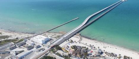 An aerial view of a coastal area with a long bridge extending into the sea, a beach with buildings and palm trees, and a clear sky.