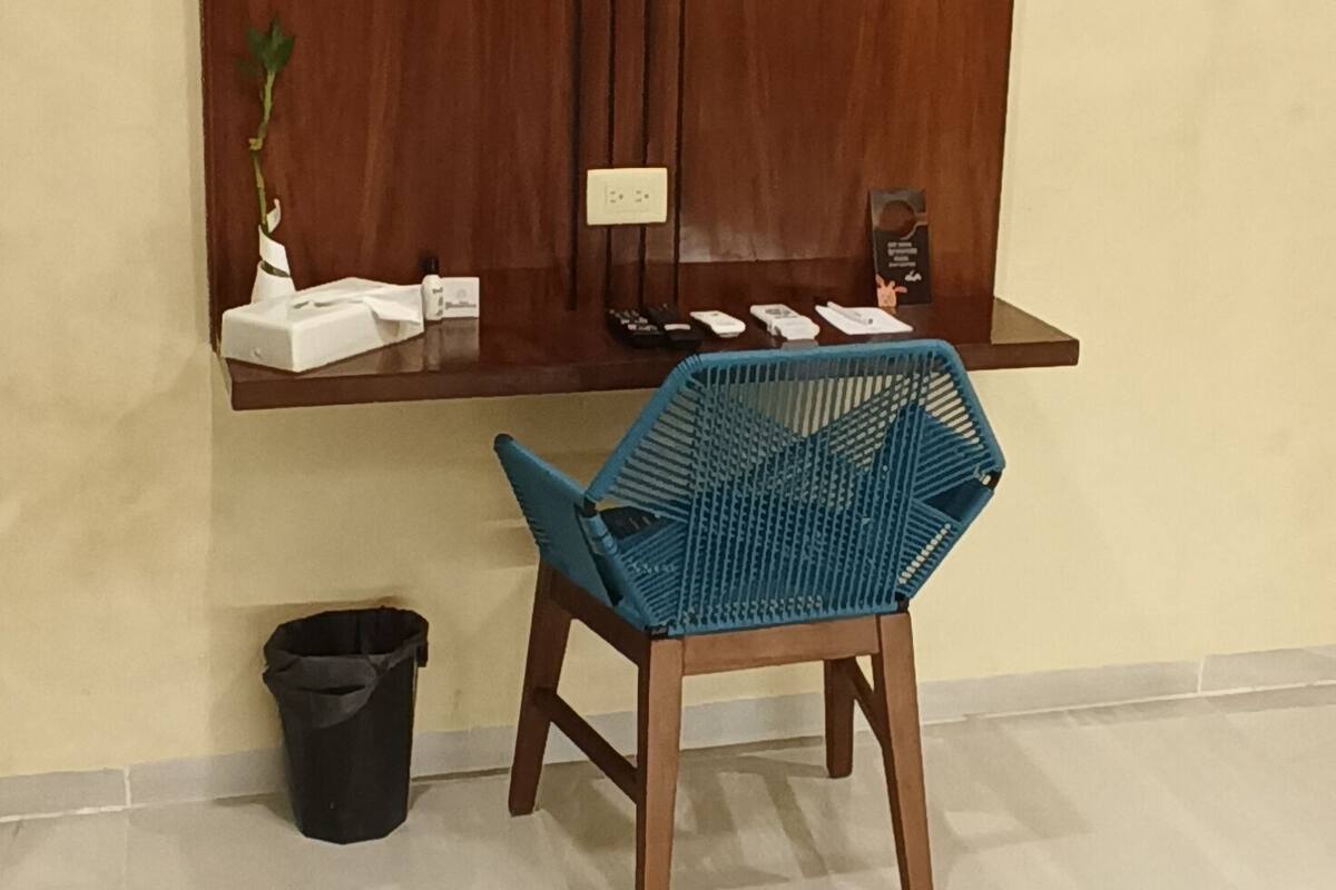 Una habitación con un televisor de pantalla plana grande montado en una pared de madera, una silla trenzada azul, un cubo de basura pequeño y una alfombra estampada en el suelo.