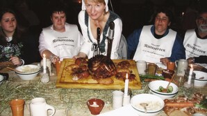 Een groep mensen rond een tafel met een gebraden varken, diverse gerechten en een kaars.