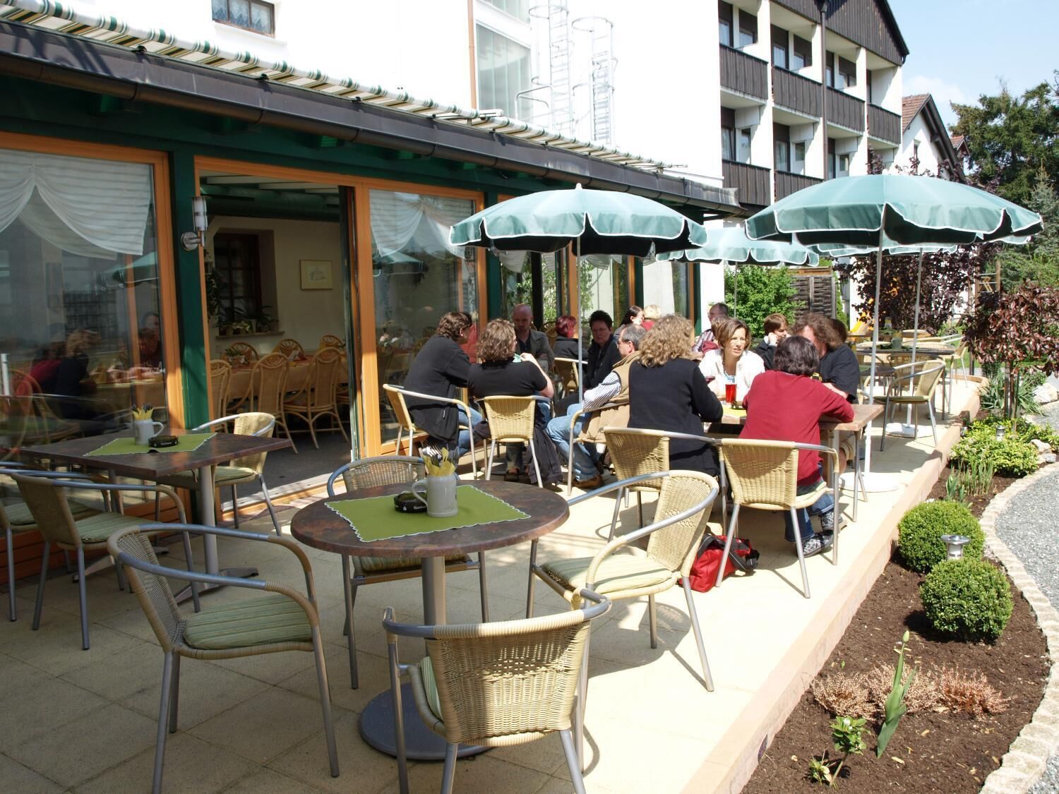 An outdoor cafe with wicker chairs and tables, umbrellas, and a building with a green awning in the background.
