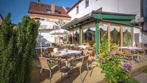 Een terras met tafels en stoelen, omgeven door groen en gebouwen.