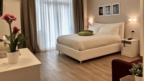 Ein modernes Hotelzimmer mit einem großen Bett, einem Nachttisch mit Lampe, einem Schreibtisch mit Blumenvase und Blick ins Freie durch transparente Vorhänge.