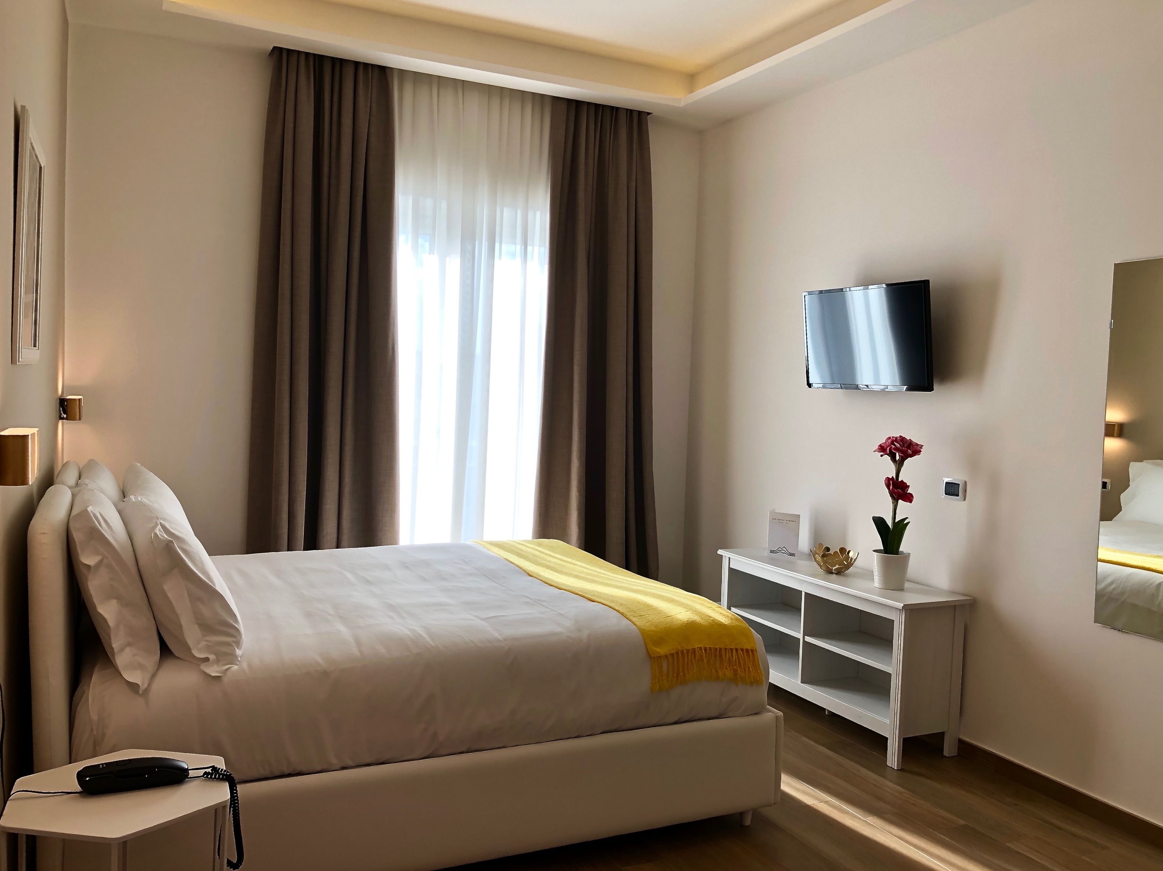 A hotel room with a bed, a bedside table with a telephone, a TV mounted on the wall, and a small table with a vase of flowers.