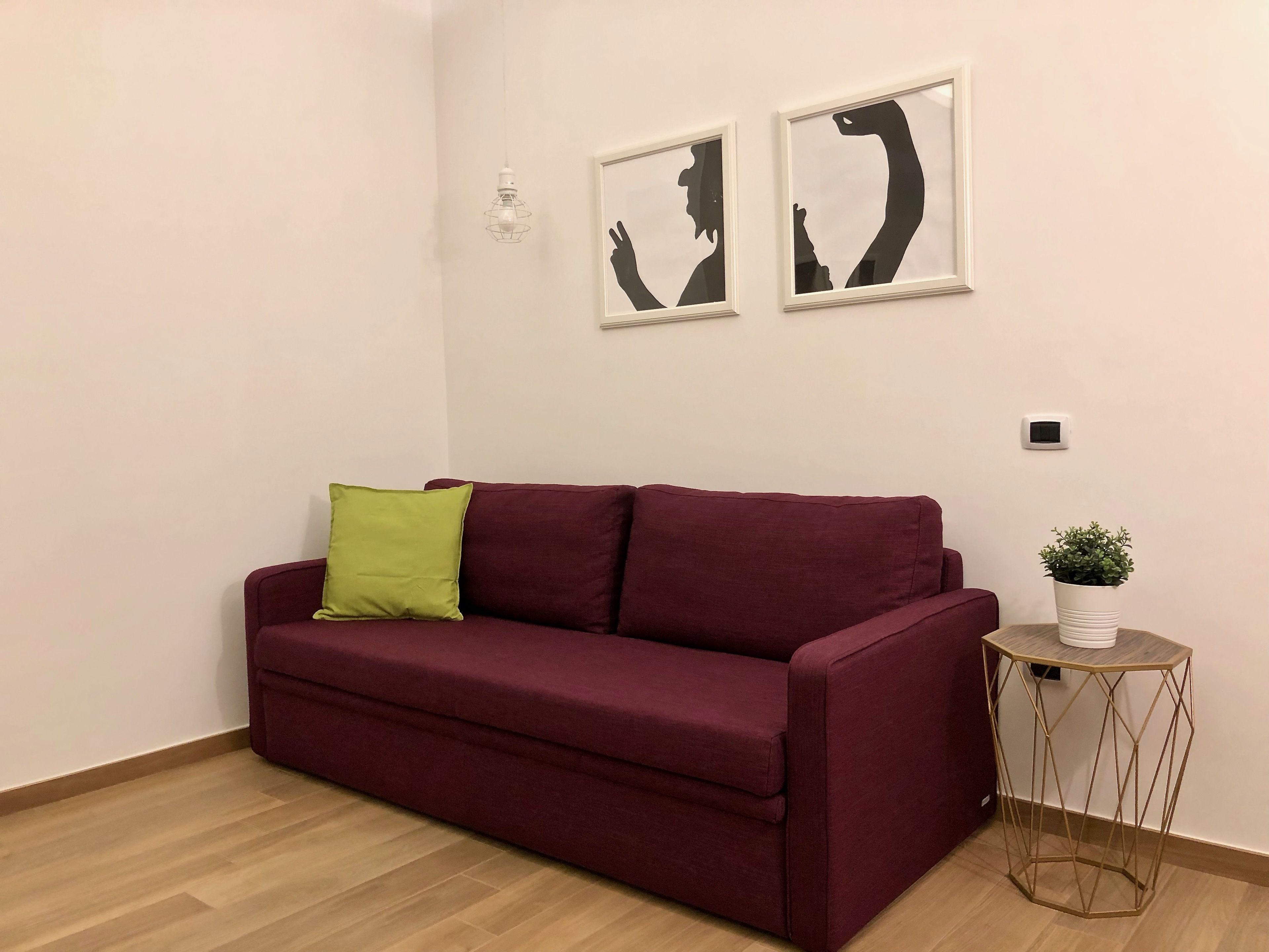 A maroon sofa with a green pillow, a small round side table with a plant, and two framed abstract wall art pieces.