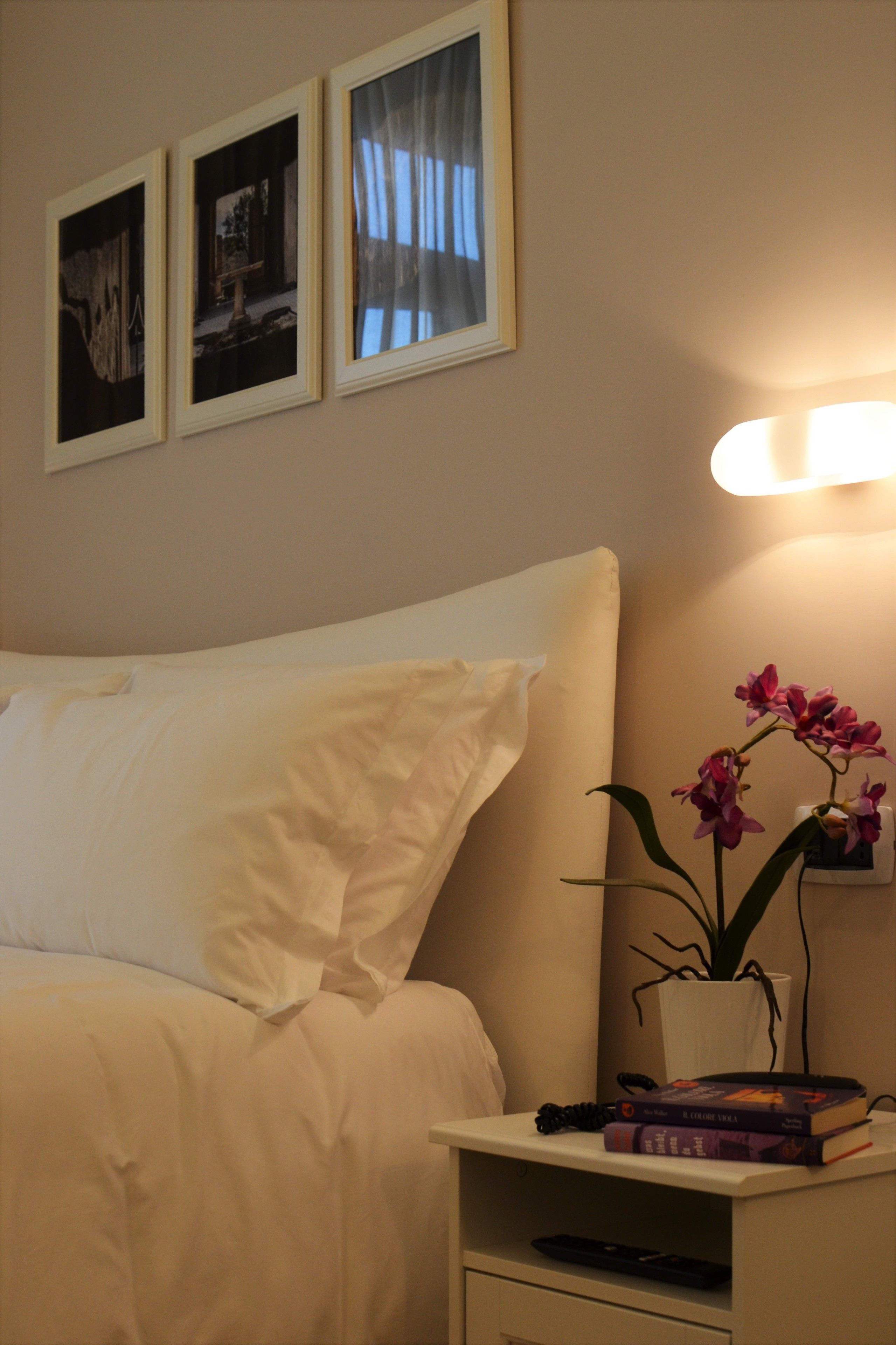 Una cama bien hecha con sábanas blancas, una mesita de noche con una lámpara, una orquídea en maceta y tres cuadros en la pared.