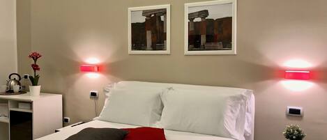 Cama bem arrumada com almofadas brancas e vermelhas, uma pequena mesa de cabeceira com uma planta em vaso e duas luzes fixadas na parede.