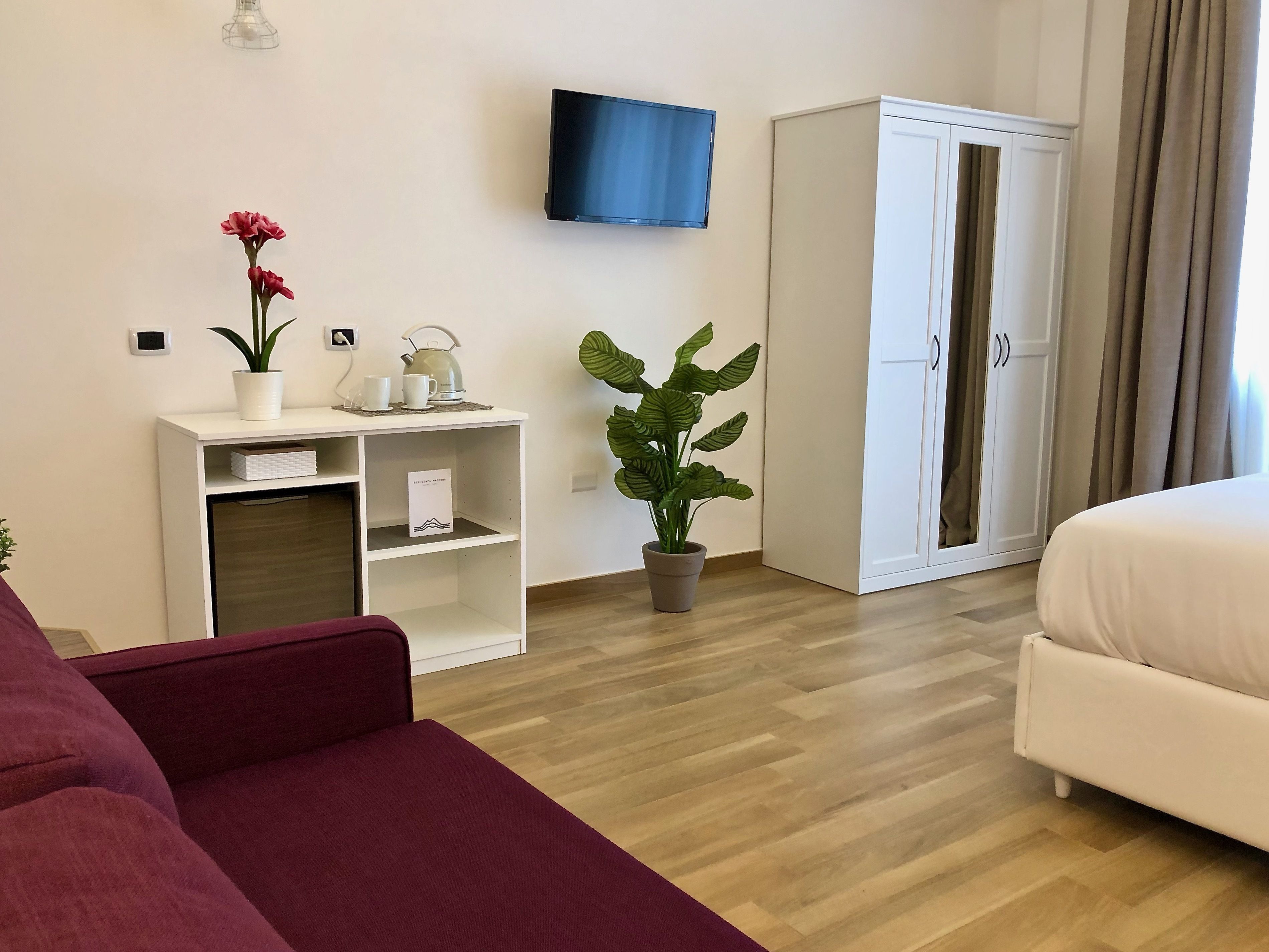 A hotel room with a bed, a TV mounted on the wall, a wardrobe, a desk with a plant and a kettle, and a red sofa.