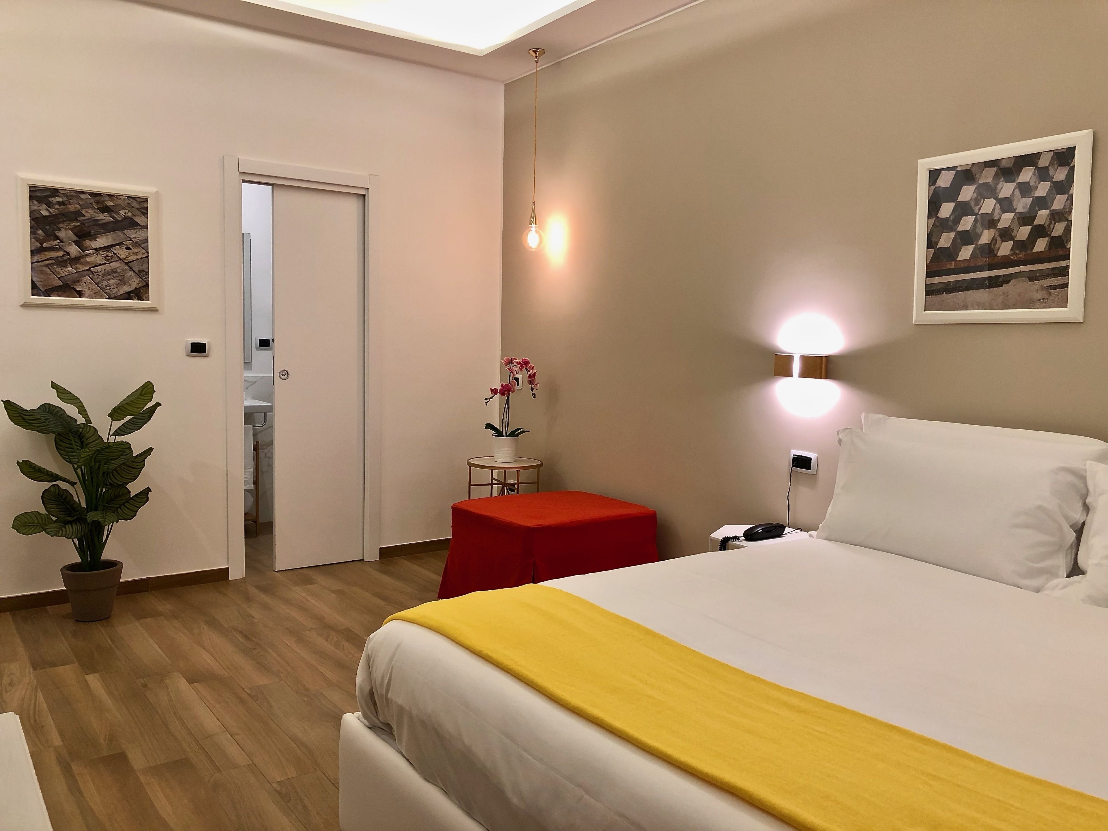 Une chambre d’hôtel avec un lit, une petite table et un tabouret rouge, une plante en pot et une lampe fixée au mur.