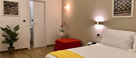 Une chambre d’hôtel avec un lit, une petite table et un tabouret rouge, une plante en pot et une lampe fixée au mur.